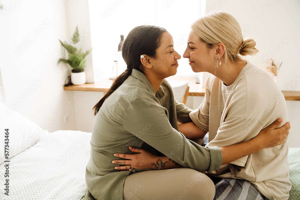 Smiling lesbian couple hugging while sitting together on bed at home