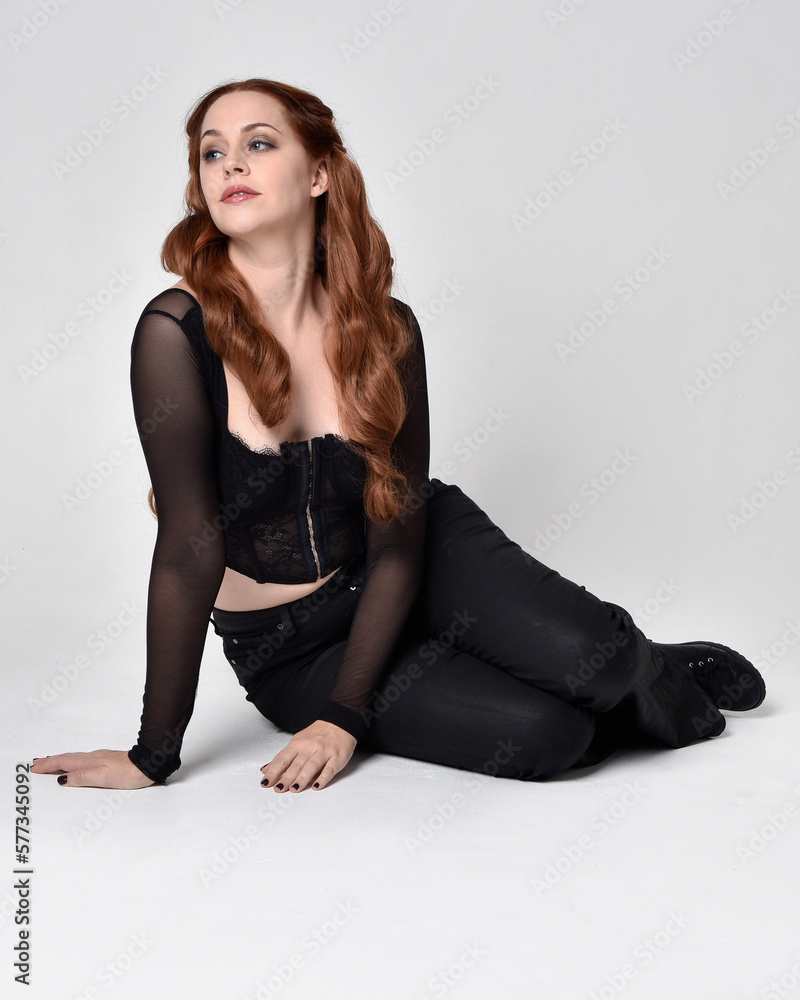 Full length portrait of beautiful woman with long red hair wearing sheer corset top and leather pants. Sitting pose with gestural hands reaching out, kneeling on floor. Isolated on white studio backg Full length portrait of beautiful woman with long red hair wearing sheer corset top and leather pants. Sitting pose with gestural hands reaching out, kneeling on floor. Isolated on white studio backg