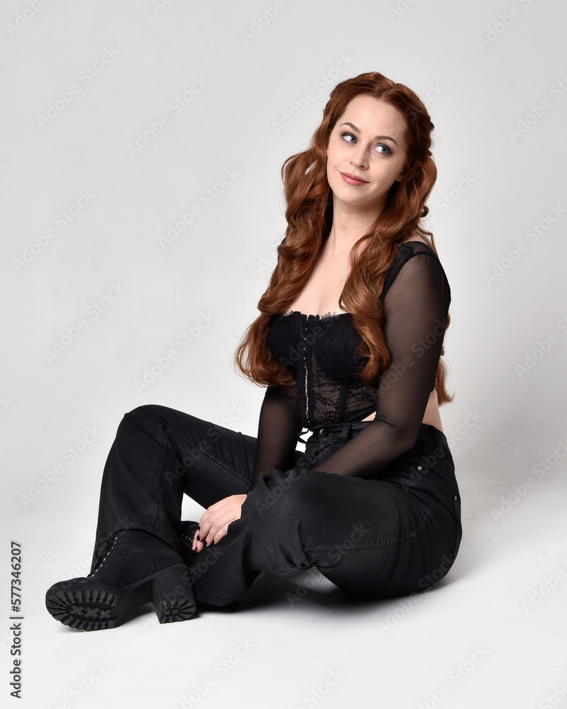 Full length portrait of beautiful woman with long red hair wearing sheer corset top and leather pants. Sitting pose with gestural hands reaching out, kneeling on floor. Isolated on white studio backg Full length portrait of beautiful woman with long red hair wearing sheer corset top and leather pants. Sitting pose with gestural hands reaching out, kneeling on floor. Isolated on white studio backg