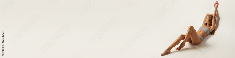 Fototapeta premium One young beautiful tanned woman with perfect figure in underwear lying on floor over gray studio background. Concept of female beauty, tenderness, body and skin care
