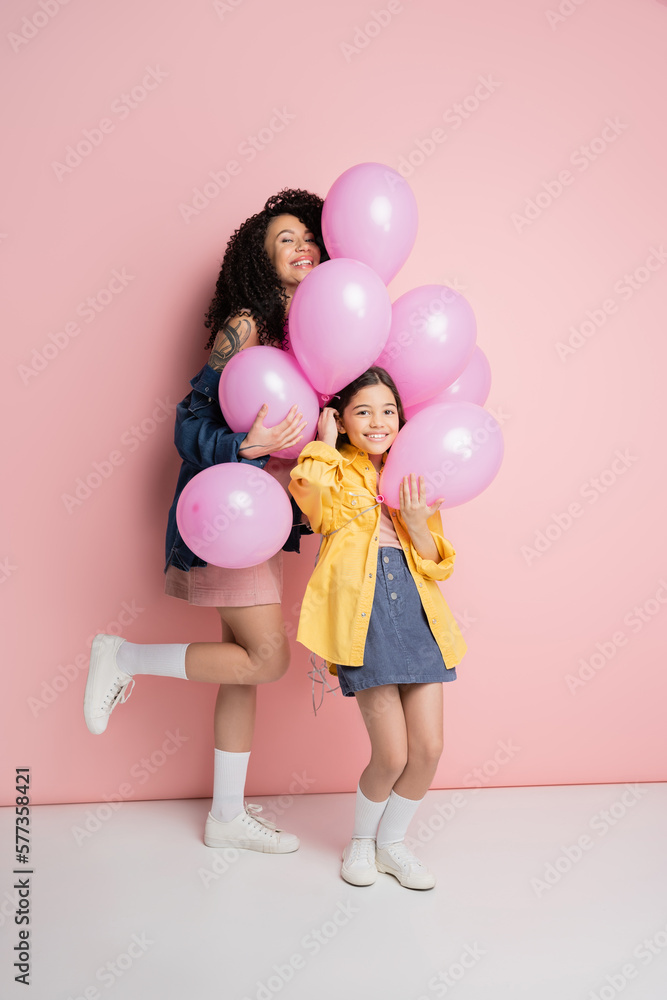 Stylish woman and daughter looking at camera near balloons on pink background. Stylish woman and daughter looking at camera near balloons on pink background.