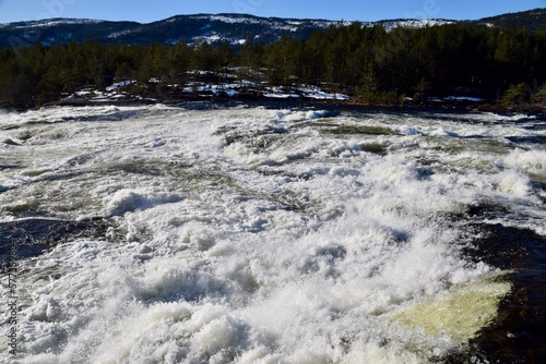 White Water rushing over rocks with Trees and hills and blue skies behind. Byglandsfjord, Norway, Feb 2023. 