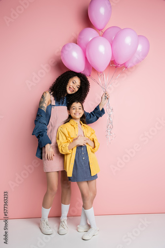 Smiling woman and kid in stylish clothes holding balloons on pink background.