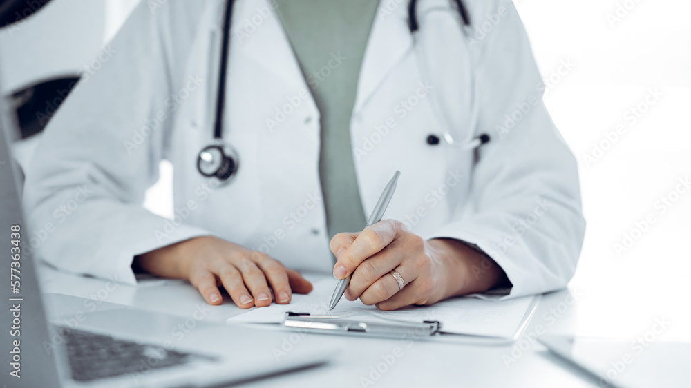 Unknown doctor woman sitting and writing notes at the desk in clinic or hospital office, close up. Medicine concept. Unknown doctor woman sitting and writing notes at the desk in clinic or hospital office, close up. Medicine concept.