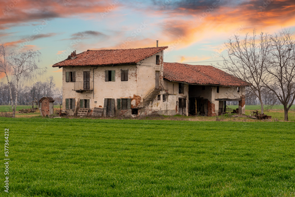 Old abandoned italian farmhouse with typical rural architecture of the Po Valley in the province of Cuneo, Italy, at sunset with field of young green wheat