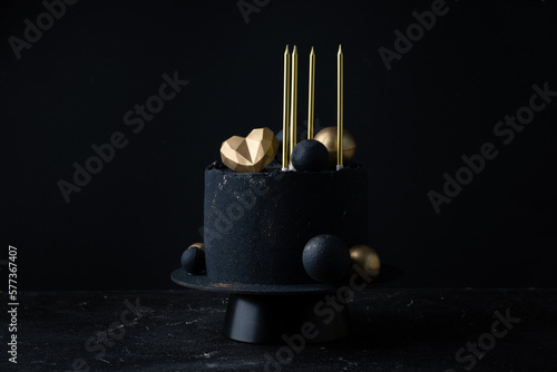 Luxury anniversary cake with black velvet sprayed coating decorated with golden chocolate spheres, heart and candles on top. Vintage cake for beloved one on the black background