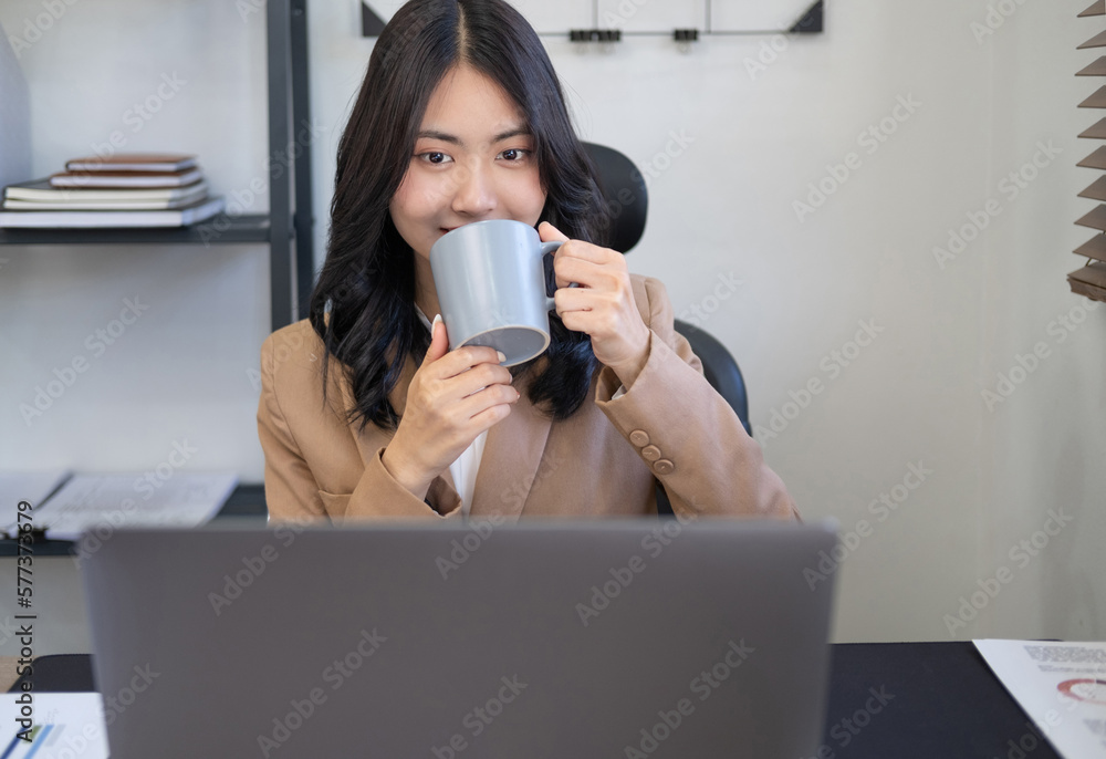 Beautiful young woman working on laptop computer while sitting at the office, drinking coffee.Friendly smile of young beautiful joyful woman front desk business woman working on her computer. Beautiful young woman working on laptop computer while sitting at the office, drinking coffee.Friendly smile of young beautiful joyful woman front desk business woman working on her computer.