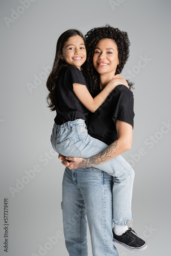 Tattooed mother lifting daughter in jeans and t-shirt isolated on grey.