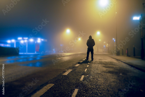 A mysterious hooded figure  back to camera. Standing on an empty road in a city. On a eerie winters night