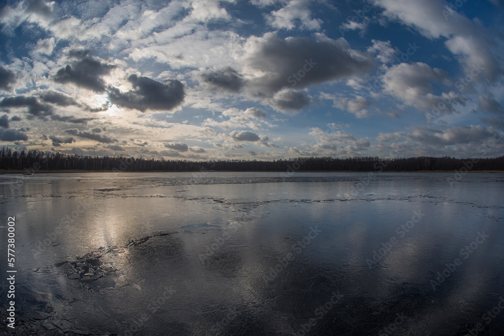 Evening landscape of a spring lake covered with thin cracking transparent ice and a cloudy sky reflected in it. Fish eye.