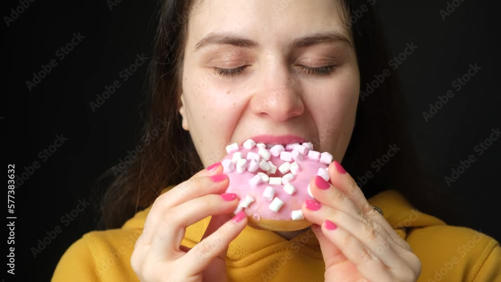 A woman licks looking at a pink marshmallow donut and bites it, a black background. A woman licks looking at a pink marshmallow donut and bites it, a black background.
