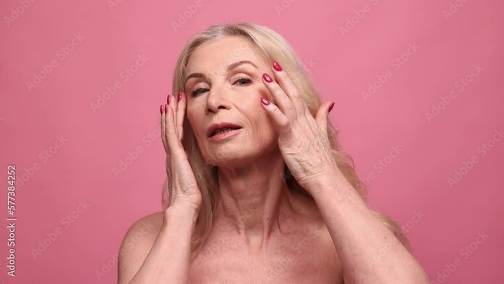 Beautiful, gorgeous middle-aged, mature woman doing face massage with fingers over pink studio background. Concept of natural beauty, face skin care, cosmetology and cosmetics, health Beautiful, gorgeous middle-aged, mature woman doing face massage with fingers over pink studio background. Concept of natural beauty, face skin care, cosmetology and cosmetics, health