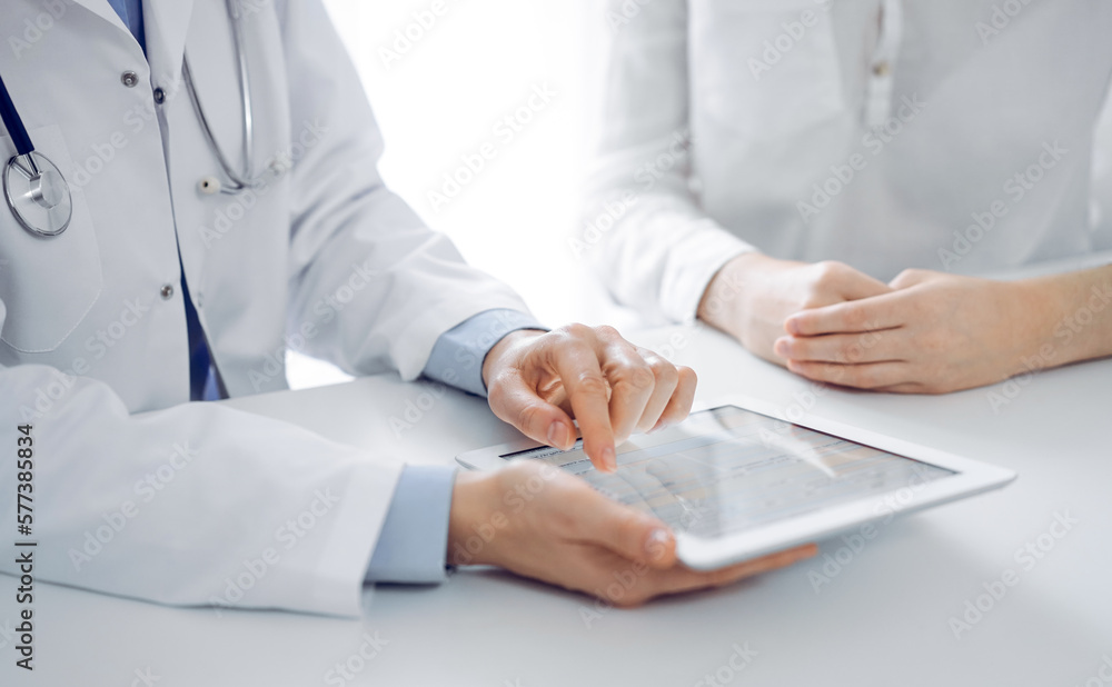 Doctor and patient sitting near each other at the desk in clinic. The focus is on female physician's hands pointing into tablet computer touchpad, close up. Medicine concept. Doctor and patient sitting near each other at the desk in clinic. The focus is on female physician's hands pointing into tablet computer touchpad, close up. Medicine concept.