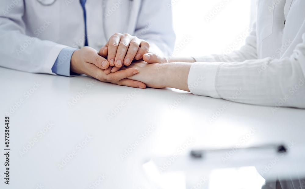 Obraz premium Doctor and patient sitting near each other at the table in clinic office. The focus is on female physician's hands reassuring woman, only hands, close up. Medicine concept.