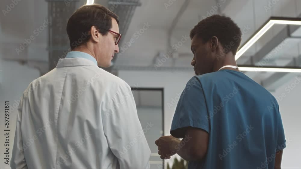Tracking medium back view of Caucasian male doctor and African American male nursing assistant having conversation while walking along hospital during shift Tracking medium back view of Caucasian male doctor and African American male nursing assistant having conversation while walking along hospital during shift