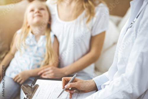 Doctor and patient. Pediatrician using clipboard while examining little girl with her mother at home. Happy cute caucasian child at medical exam. Medicine concept.