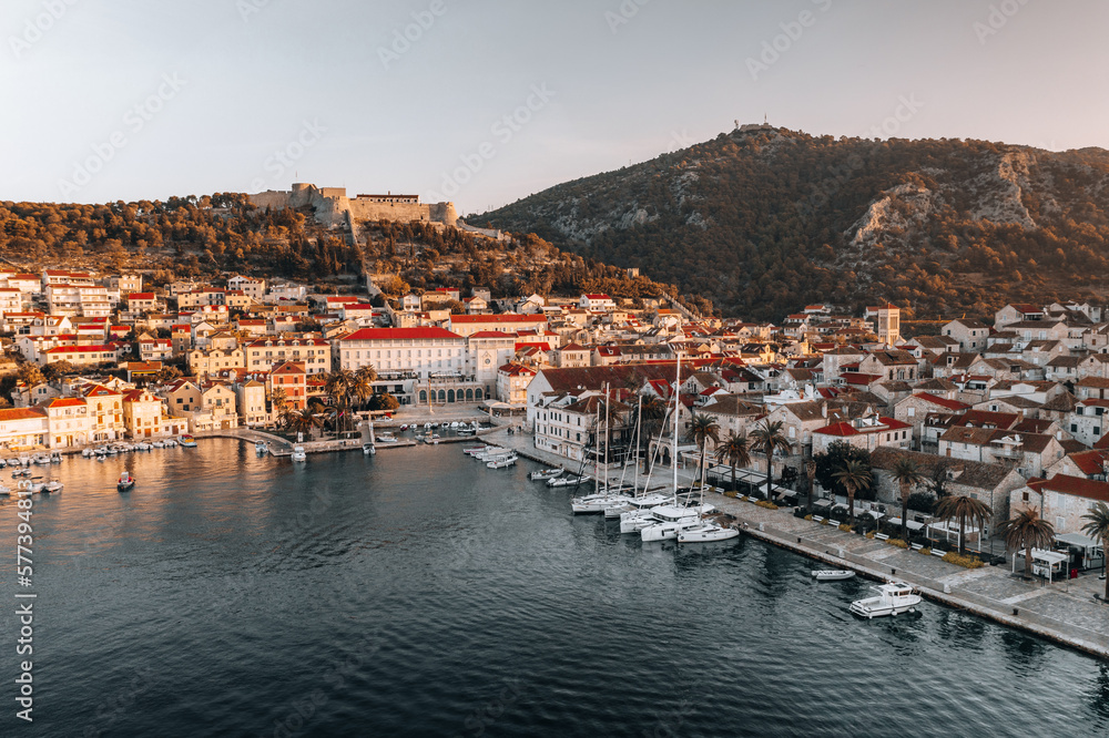 Aerial shot captures the beauty of Hvar's coastline at sunrise on a summer morning. The crystal-clear waters and rugged cliffs are illuminated by the soft, warm light of the rising sun