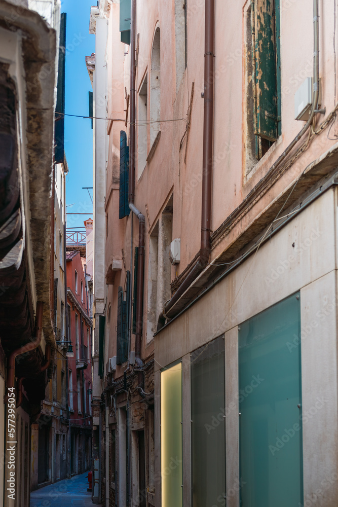 View of a narrow street in Venice with a strip of blue sky in the background on a sunny day, old vintage walls of authentic houses, Italian medieval architecture, Venice culture, tourist attractions