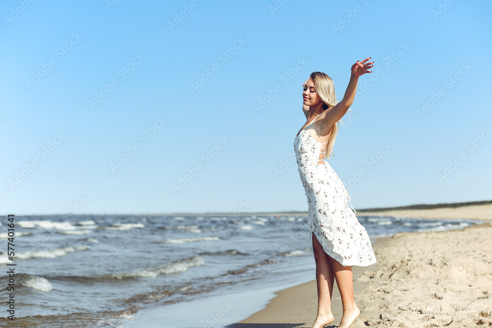 Happy blonde beautiful woman on the ocean beach standing in a white summer dress, open arms. Happy blonde beautiful woman on the ocean beach standing in a white summer dress, open arms.