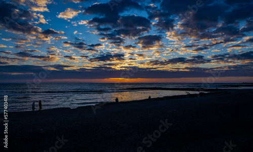Some people take a walk on the beach of Ostia, Rome, in the sunset, in winter. The sun covered by clouds and the reflection of warm light on the blue sea water.