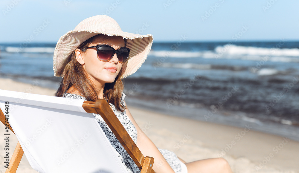 Happy brunette woman wearing sunglasses and hat relaxing on a wooden deck chair at the ocean beach.