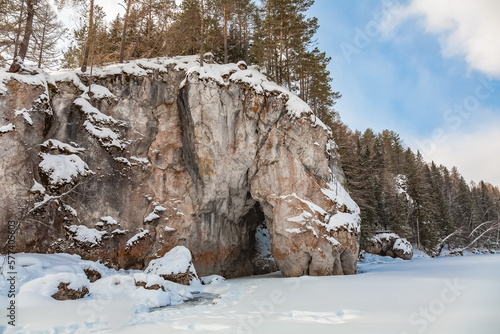 Winter landscape with a rock in the form of a horse 's head drinking water and trees on the bank of a snow - covered river against a cloudy blue sky