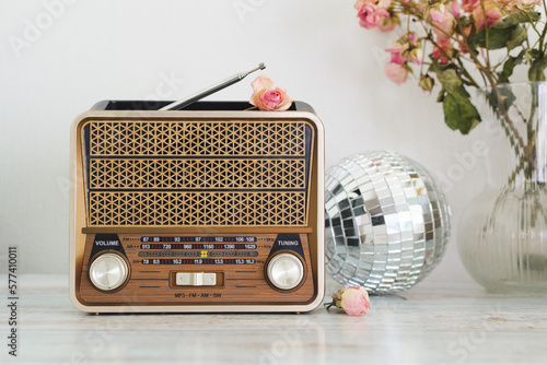 On the table against the background of a light wall is a retro radio behind which is a disco ball and a vase with a dried bouquet of pink roses, copy space.