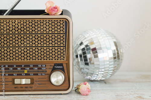 Close-up of a vintage retro radio against a light wall on a wooden table, in the background a glass disco ball and buds of dried pink roses, copy space.