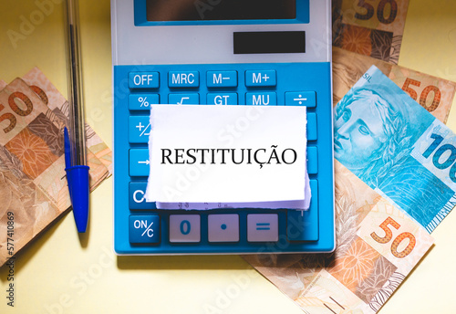 A palavra Restituição em Português do Brasil escrito em um pedaço de papel que está sobre uma calculadora. Notas do Real Brasileiro na composição. Economia Brasileira. photo