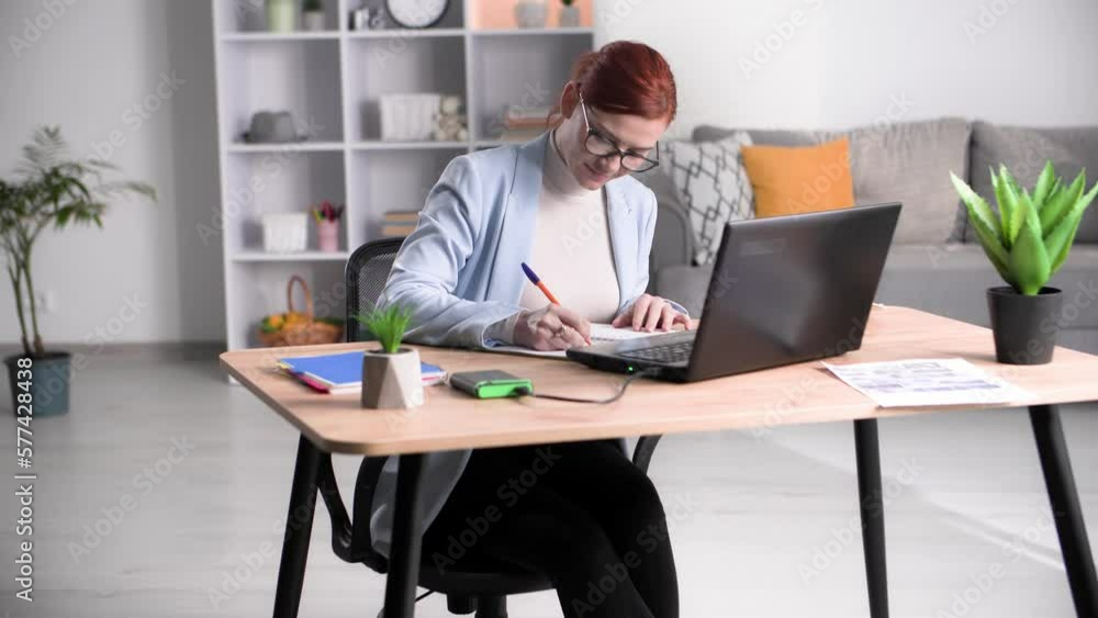 online learning, charming woman in glasses making notes in a notebook and typing on laptop while sitting at home online learning, charming woman in glasses making notes in a notebook and typing on laptop while sitting at home