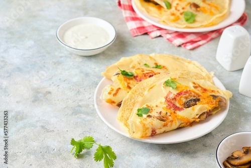 Hearty wheat pancakes with ham, mushrooms and cheddar cheese on a white plate on a gray concrete background. International Pancake Day, Maslenitsa.