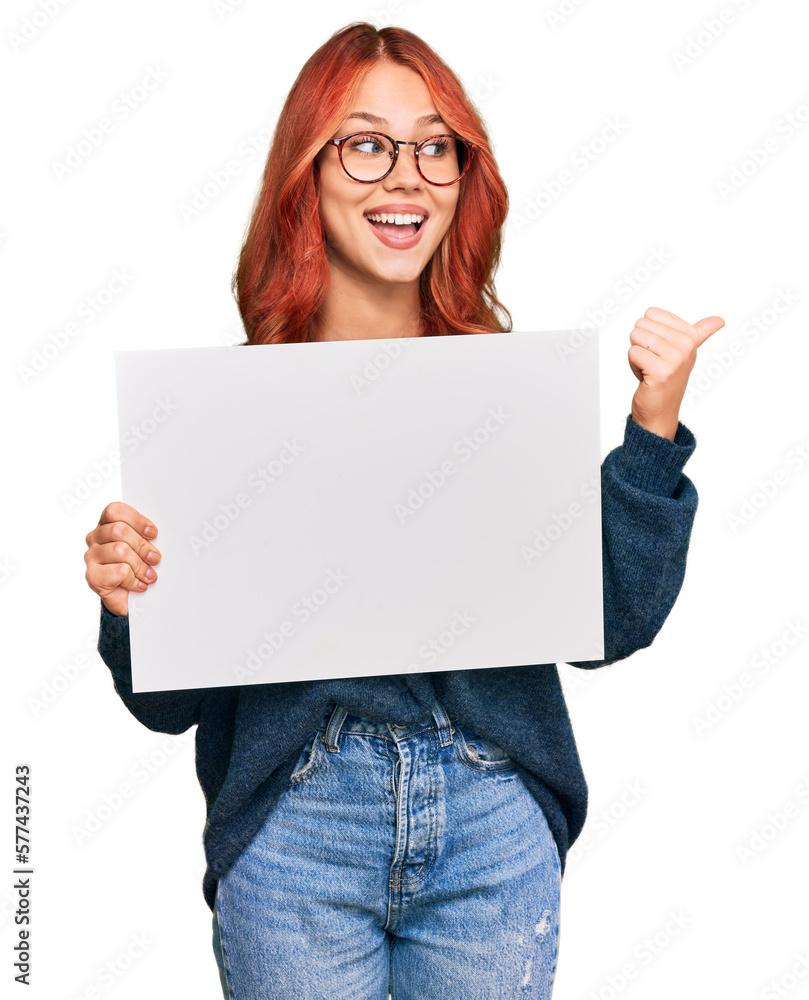 Young redhead woman holding blank empty banner pointing thumb up to the side smiling happy with open mouth Young redhead woman holding blank empty banner pointing thumb up to the side smiling happy with open mouth