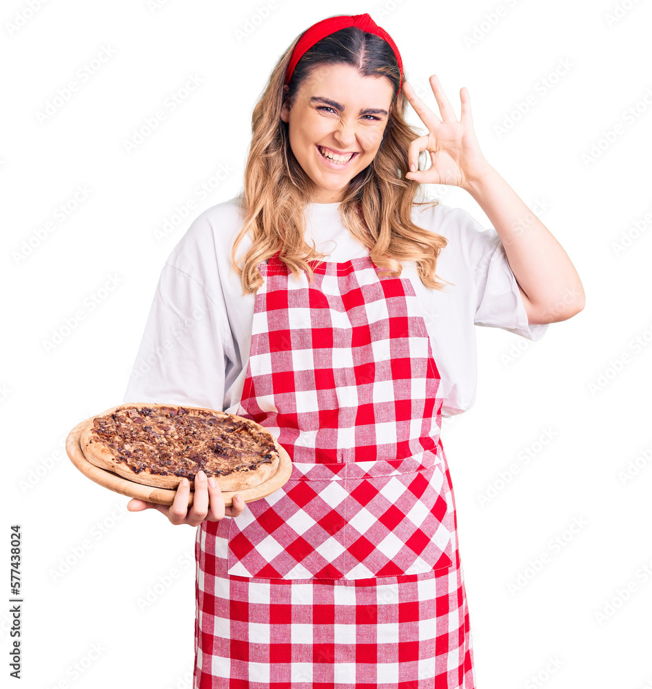 Young caucasian woman wearing apron holding pizza doing ok sign with fingers, smiling friendly gesturing excellent symbol Young caucasian woman wearing apron holding pizza doing ok sign with fingers, smiling friendly gesturing excellent symbol