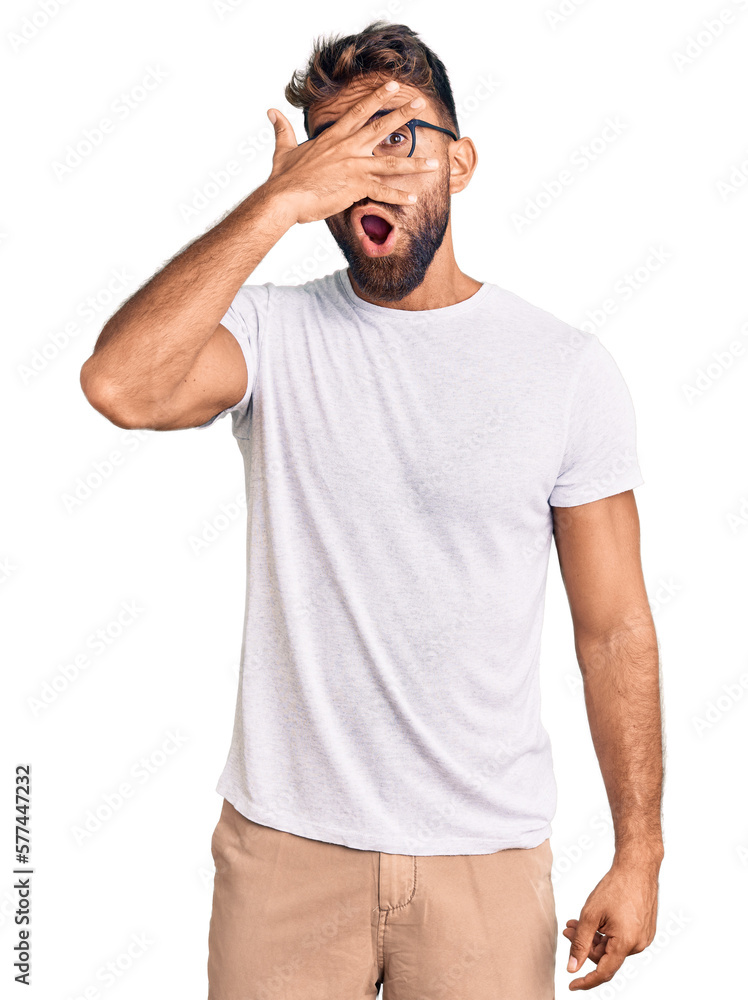 Young hispanic man wearing casual clothes and glasses peeking in shock covering face and eyes with hand, looking through fingers with embarrassed expression. Young hispanic man wearing casual clothes and glasses peeking in shock covering face and eyes with hand, looking through fingers with embarrassed expression.