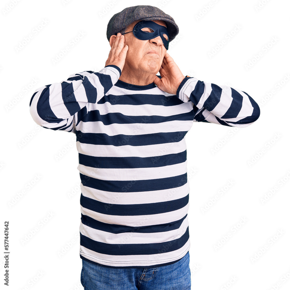 Senior handsome man wearing burglar mask and t-shirt covering ears with fingers with annoyed expression for the noise of loud music. deaf concept. Senior handsome man wearing burglar mask and t-shirt covering ears with fingers with annoyed expression for the noise of loud music. deaf concept.