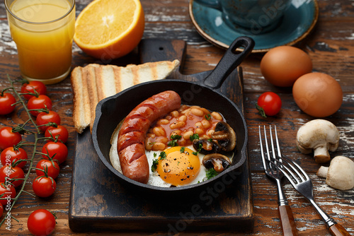 Full english breakfast with fried eggs, grilled sausage, beans, mushrooms, toasts, cherry tomatoes, orange juice and cup of coffee on wooden rustic background. Side view, close up
