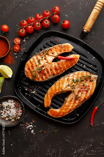 Two grilled salmon steaks in cast-iron pan with vegetables and spices on dark concrete background. Top view, flat lay