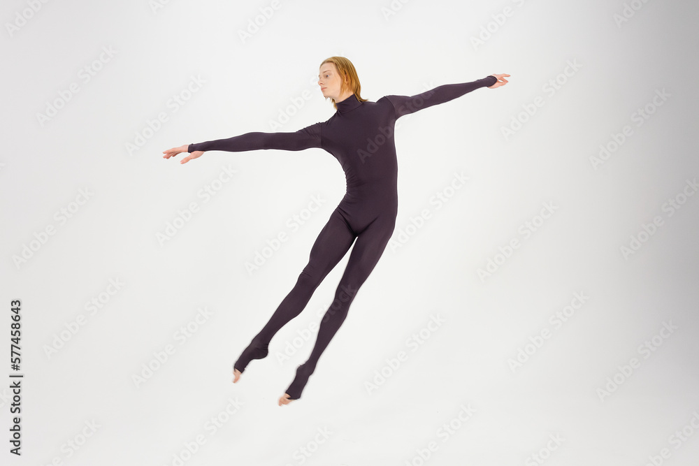 Young athletic professional ballet dancer in a black unitard is in perfect shape, performing and jumping over a white background.