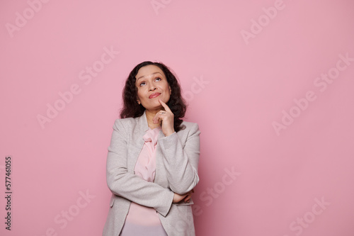 Pensive middle-aged woman in elegant light gray suit, thoughtfully looking up, holding finger at her chin, reasoning, finding solutions of problems, isolated pink color background with copy ad space