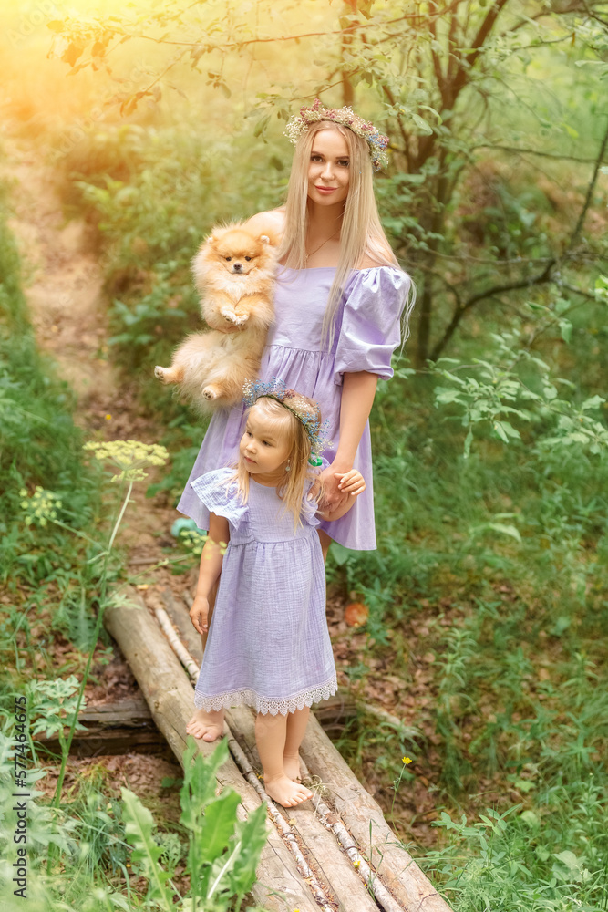 Mom and daughter, dressed in lilac dresses and wreaths of flowers, stand on a wooden bridge in the summer forest. Pomeranian dog in woman's hands Mom and daughter, dressed in lilac dresses and wreaths of flowers, stand on a wooden bridge in the summer forest. Pomeranian dog in woman's hands