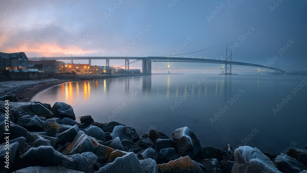 Two bridges covered with thick fog at sunset in winter and a stone embankment in hoarfrost. Forth Road Bridge and Queensferry Crossing. South Queensferry, Scotland, United Kingdom