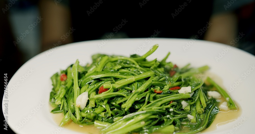 Close up person holding dish of cooked healthy vegetable stir fried fresh green Spicy water spinach seasoned with spicy chili and soy sauce , Thai food, fresh morning glory with red chili and garlic
