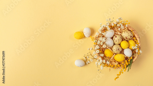 Easter candy chocolate eggs and almond sweets lying in a bird's nest decorated with flowers and feathers on a yellow background. Top view long banner. Happy Easter concept.