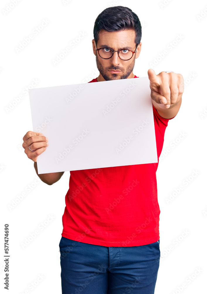 Young hispanic man holding blank empty banner pointing with finger to the camera and to you, confident gesture looking serious Young hispanic man holding blank empty banner pointing with finger to the camera and to you, confident gesture looking serious