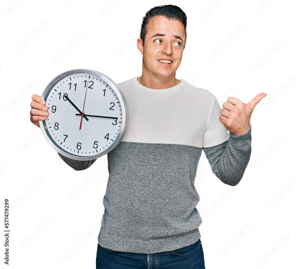 Handsome young man holding big clock pointing thumb up to the side smiling happy with open mouth Handsome young man holding big clock pointing thumb up to the side smiling happy with open mouth