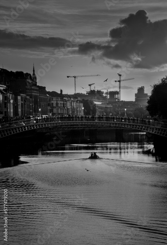 Black and white shot of a cloudy sunset over River Liffey. Silhouettes of people crossing a bridge over the water, a boat crossing under the bridge, and seagulls flying in the cityscape of Dublin