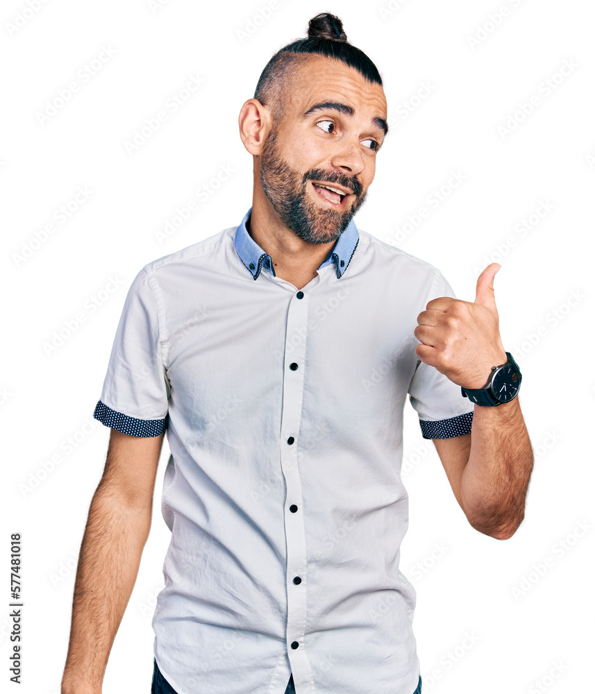 Hispanic man with ponytail wearing casual white shirt smiling with happy face looking and pointing to the side with thumb up. Hispanic man with ponytail wearing casual white shirt smiling with happy face looking and pointing to the side with thumb up.
