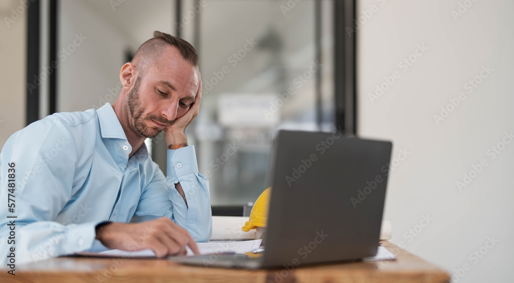Bored and tired caucasian male engineer or architect sits at his desk, hands on his chin, looking at the camera, feeling no energy to work, overworked, and lacking passion. Bored and tired caucasian male engineer or architect sits at his desk, hands on his chin, looking at the camera, feeling no energy to work, overworked, and lacking passion.
