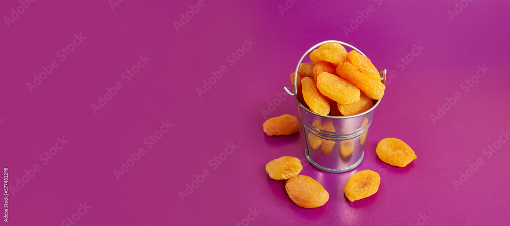 A small metal bucket filled with dried apricots on a pink background. Dried apricots as an ingredient. A small metal bucket filled with dried apricots on a pink background. Dried apricots as an ingredient.