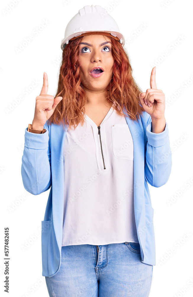 Young latin woman wearing architect hardhat amazed and surprised looking up and pointing with fingers and raised arms. Young latin woman wearing architect hardhat amazed and surprised looking up and pointing with fingers and raised arms.